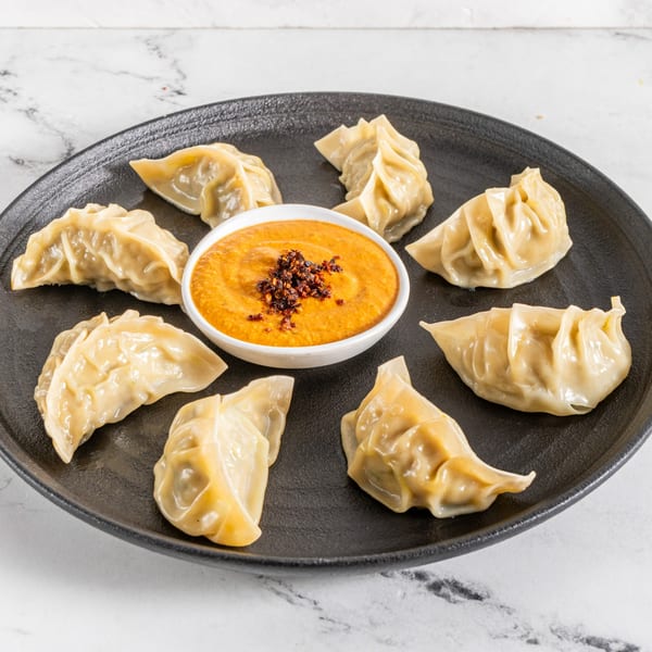 Seven steamed momos arranged around a bowl of spicy orange dipping sauce on a black plate.