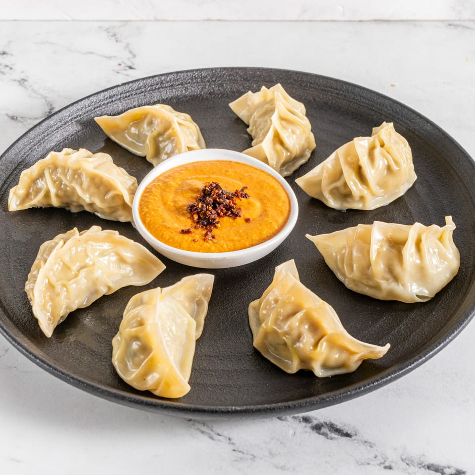 Seven steamed momos arranged around a bowl of spicy orange dipping sauce on a black plate.