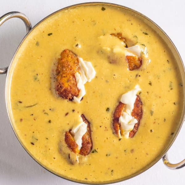 Indian Malai Kofta in a creamy yellow sauce, garnished with white cream in a metal bowl.