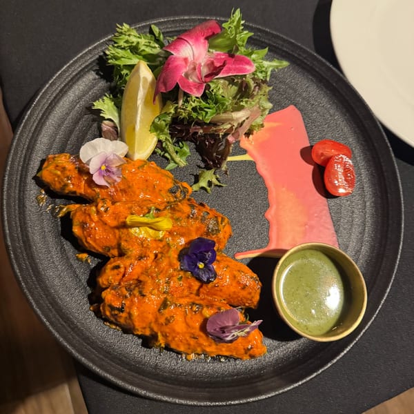 Orange chicken pieces with edible flowers, salad, lemon, and dipping sauces on a black plate.