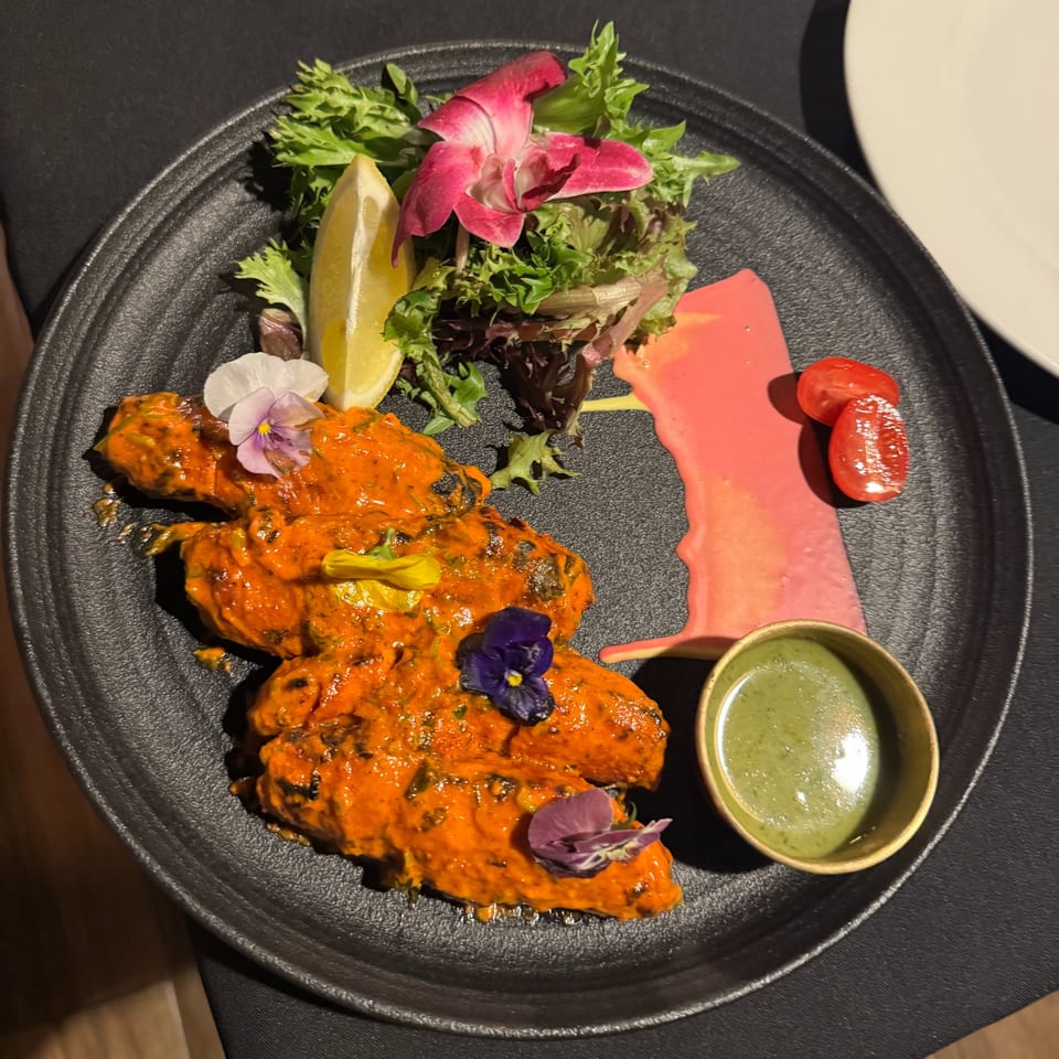 Orange chicken pieces with edible flowers, salad, lemon, and dipping sauces on a black plate.