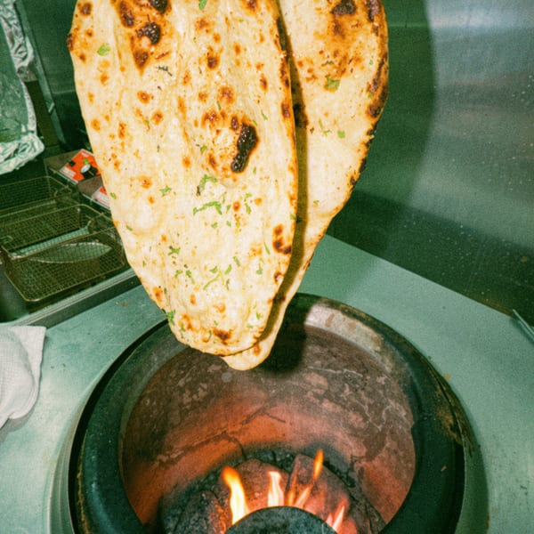 Freshly baked naan bread, garnished with herbs, being removed from a fiery tandoor oven.