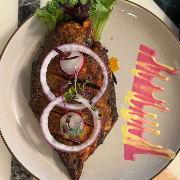 Indian-spiced whole fish, garnished with red onion, radish, and microgreens, on a plate with pink and yellow sauces.