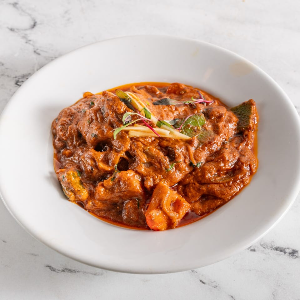 A white bowl filled with a rich, reddish-brown Indian curry, garnished with fresh herbs and ginger.