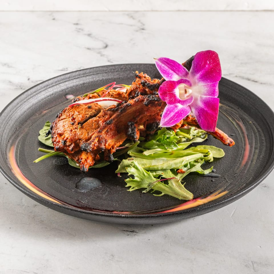 Indian lamb chops, garnished with a purple orchid and fresh greens, on a black plate.