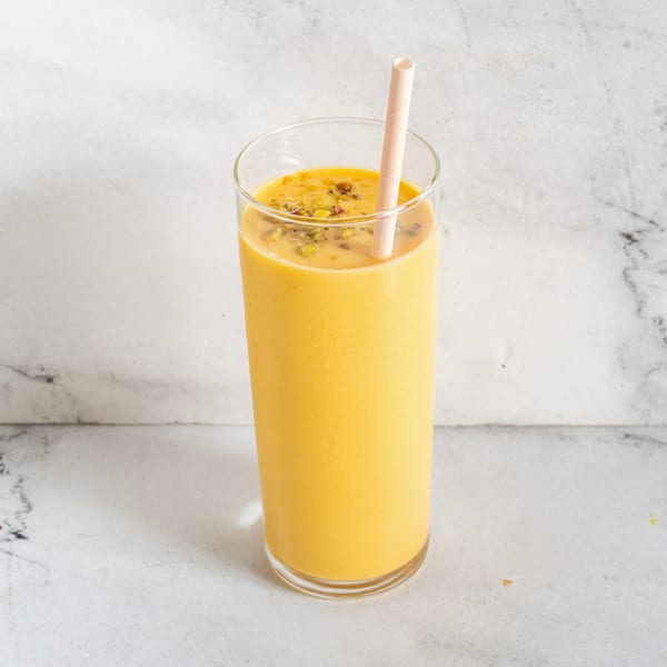 A tall glass of yellow mango lassi, topped with spices and a straw, on a white marble surface.
