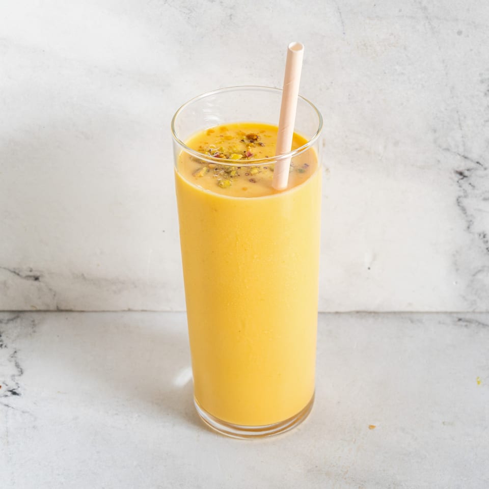 A tall glass of yellow mango lassi, topped with spices and a straw, on a white marble surface.
